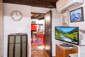 A room with a television displaying a mountainous landscape, a clock on the wall, and a door leading to a kitchen area.