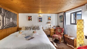 A bedroom with a wooden ceiling, a large bed with white bedding, a wooden nightstand, a chair, a lamp, and framed pictures on the wall.