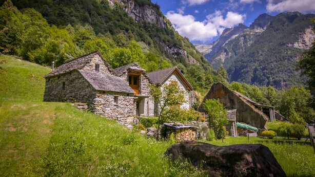 A stone cottage with a thatched roof, surrounded by lush greenery and mountains in the background.