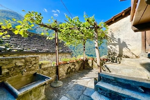 Un patio con paredes de piedra, un lavamanos de piedra, escalones de madera y una pérgola cubierta de plantas trepadoras.