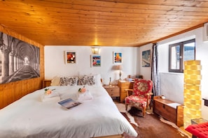 A hotel room with a wooden ceiling, a bed with white linens, a floral armchair, a desk with a lamp, and a large window with a view.