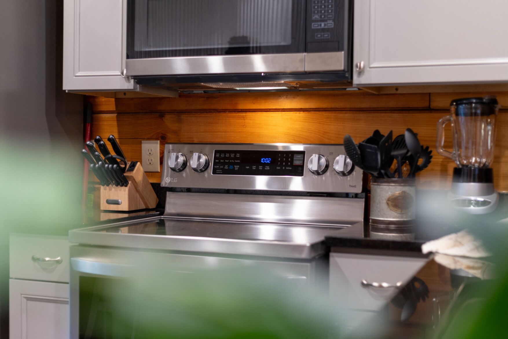A kitchen with a stainless steel stove, microwave, and blender. There are wooden cabinets and a knife block visible.