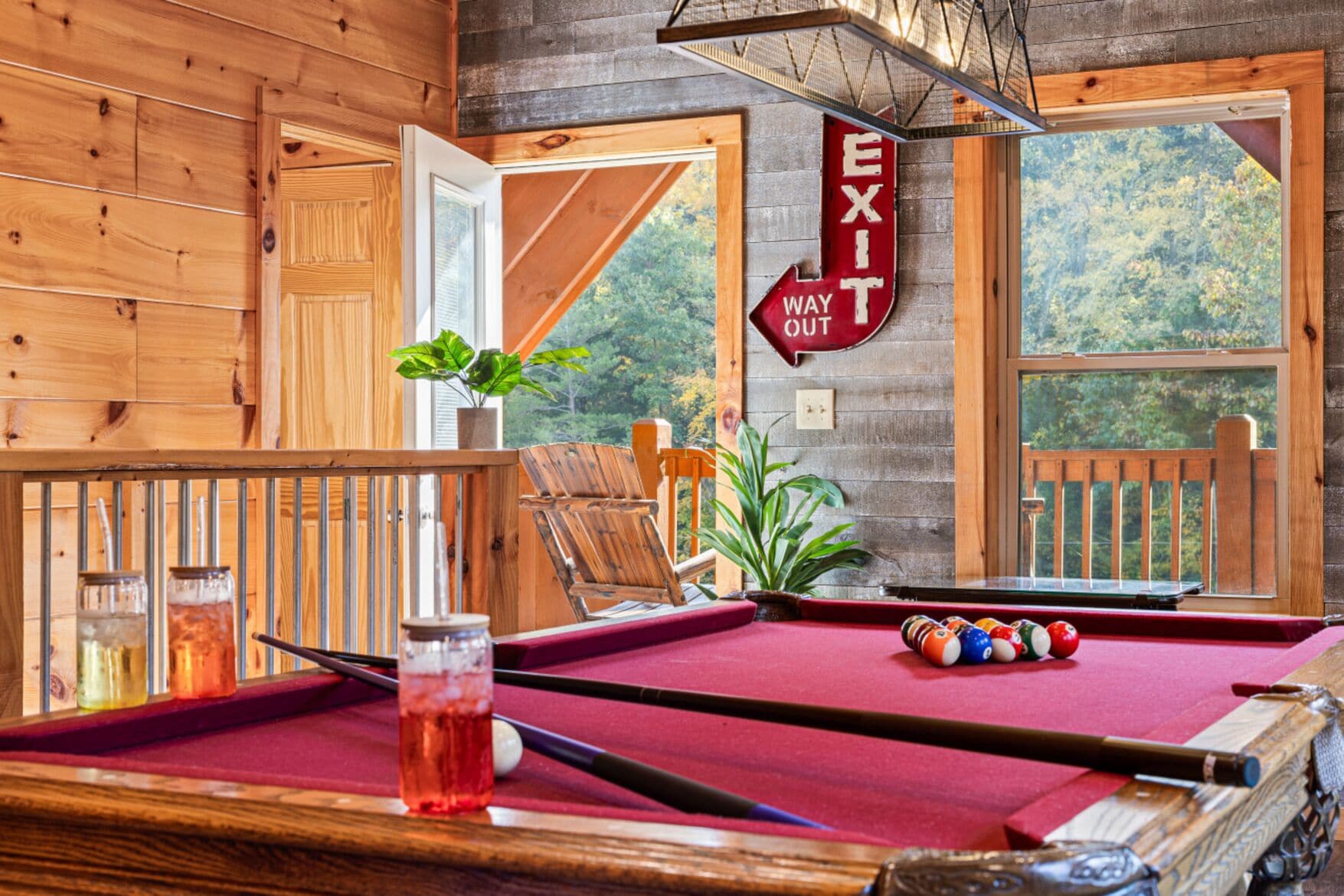 Una mesa de billar con superficie de fieltro rojo, una vara de billar y bolas de billar en una habitación con paredes de madera y vistas a árboles a través de una ventana.