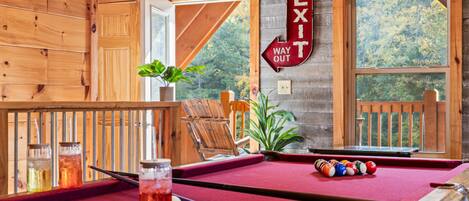 Una mesa de billar con superficie de fieltro rojo, una vara de billar y bolas de billar en una habitación con paredes de madera y vistas a árboles a través de una ventana.