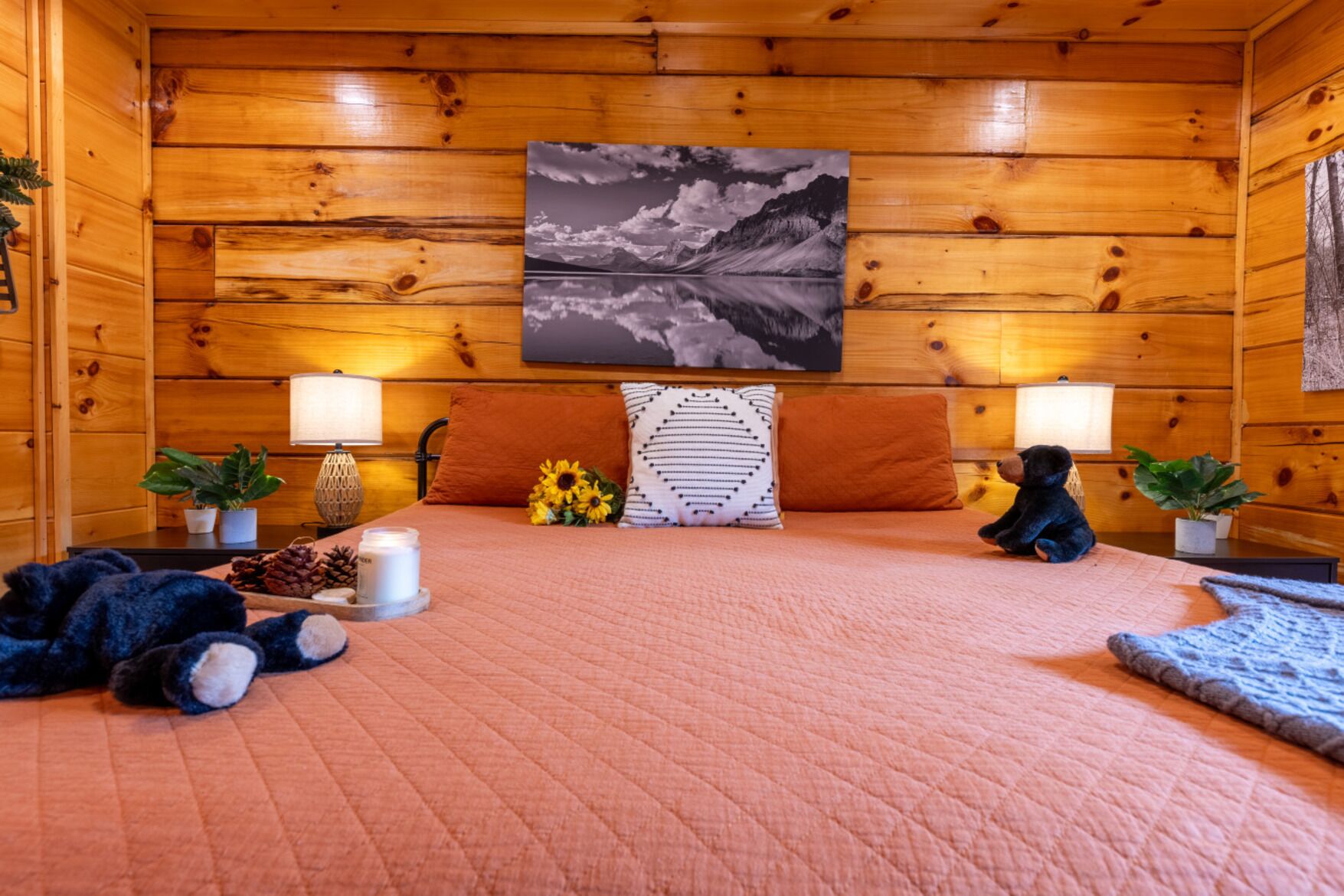 A cozy log cabin room with a bed, two nightstands, a lamp, a painting, and a teddy bear.