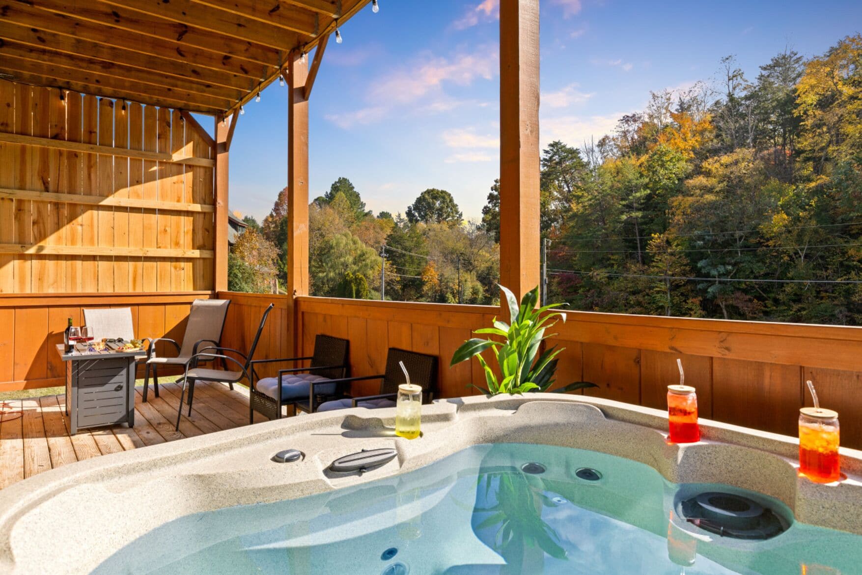 Un bain à remous avec des boissons, une table et des chaises sur une terrasse en bois, offrant une vue sur les arbres.