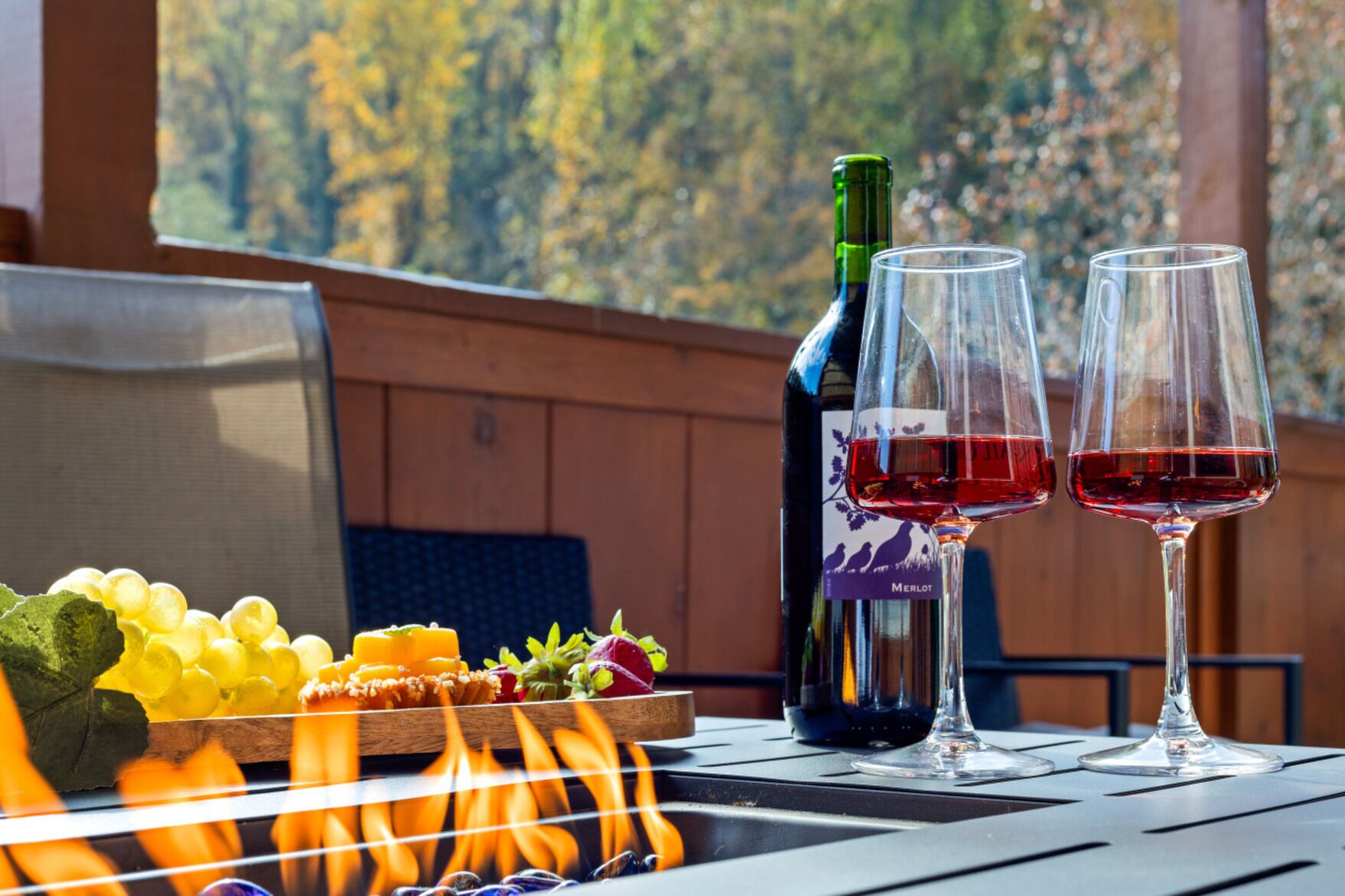 Una botella de vino Merlot, dos copas de vino tinto, un plato de frutas y una parrilla con llamas.