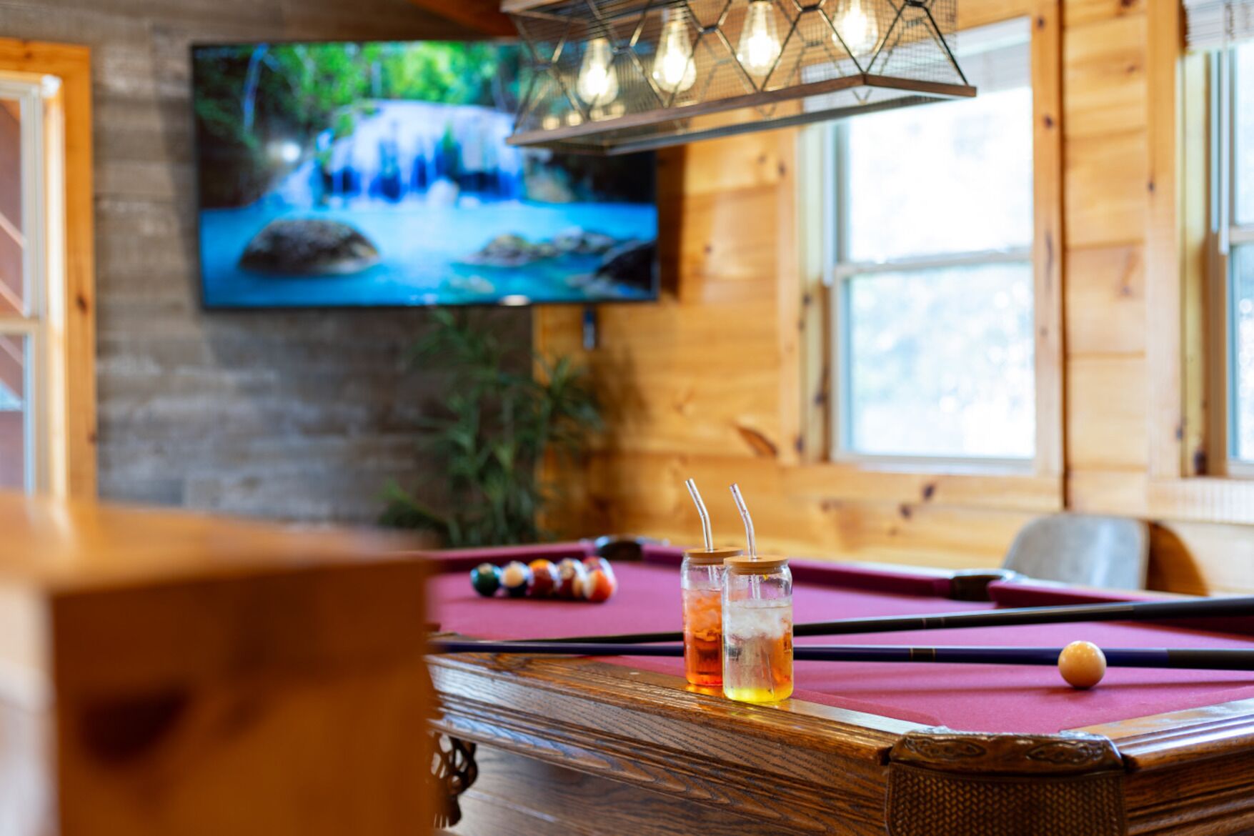 A pool table with a red felt surface, two shot glasses with straws, and a cue ball. A television mounted on a brick wall displays a waterfall scene.