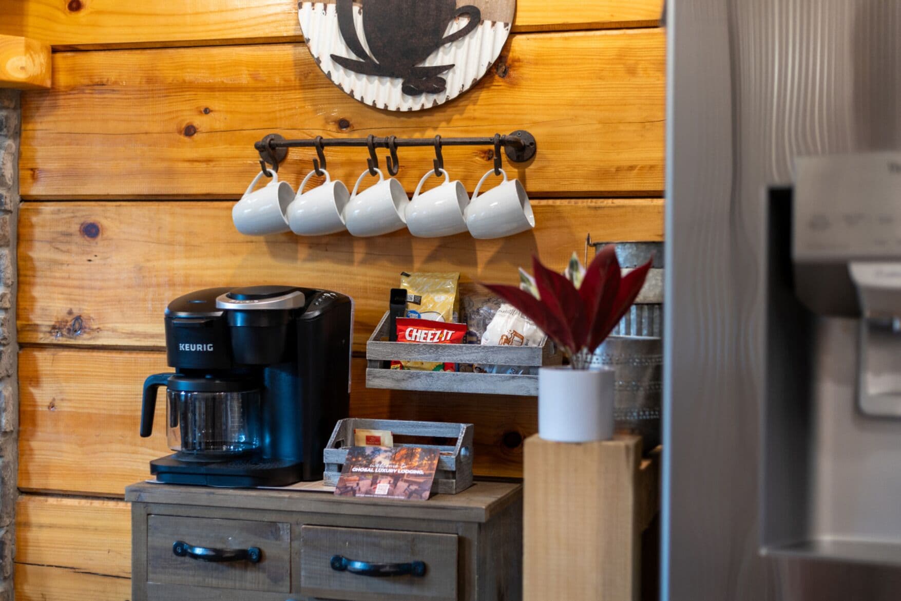 Una kitchenette con una cafetera Keurig, un estante con tazas de café, una caja de galletas Cheez-It y una pequeña planta.