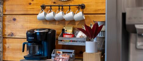 Una kitchenette con una cafetera Keurig, un estante con tazas de café, una caja de galletas Cheez-It y una pequeña planta.
