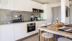 A modern kitchen with white cabinetry, a built-in oven, a coffee machine, and a dining area with a wooden table and chairs.