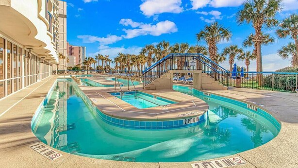 A pool area with multiple swimming pools, palm trees, and a building with balconies. - Direct Oceanfront Two Bedroom Condo (Myrtle Beach)