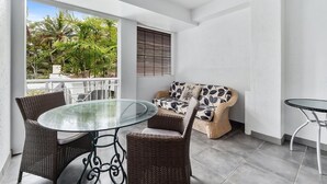 A balcony with a glass table, wicker chairs, a sofa with patterned cushions, and a ceiling fan. - Alassio 1 Bedroom Pool View Apartment 204/1 (Cairns)