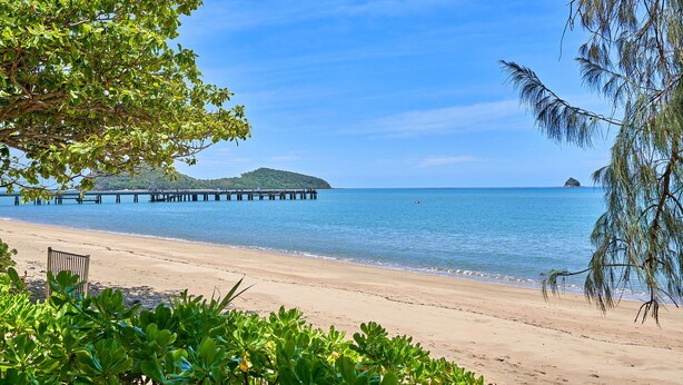A beach with clear blue water, a pier, and a distant island. - Alassio 1 Bedroom Pool View Apartment 204/1 (Cairns)