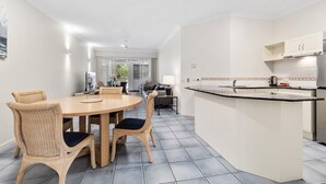 A modern kitchen with a central island, dining area with wicker chairs, and a view of the living room. - Alassio 1 Bedroom Pool View Apartment 204/1 (Cairns)