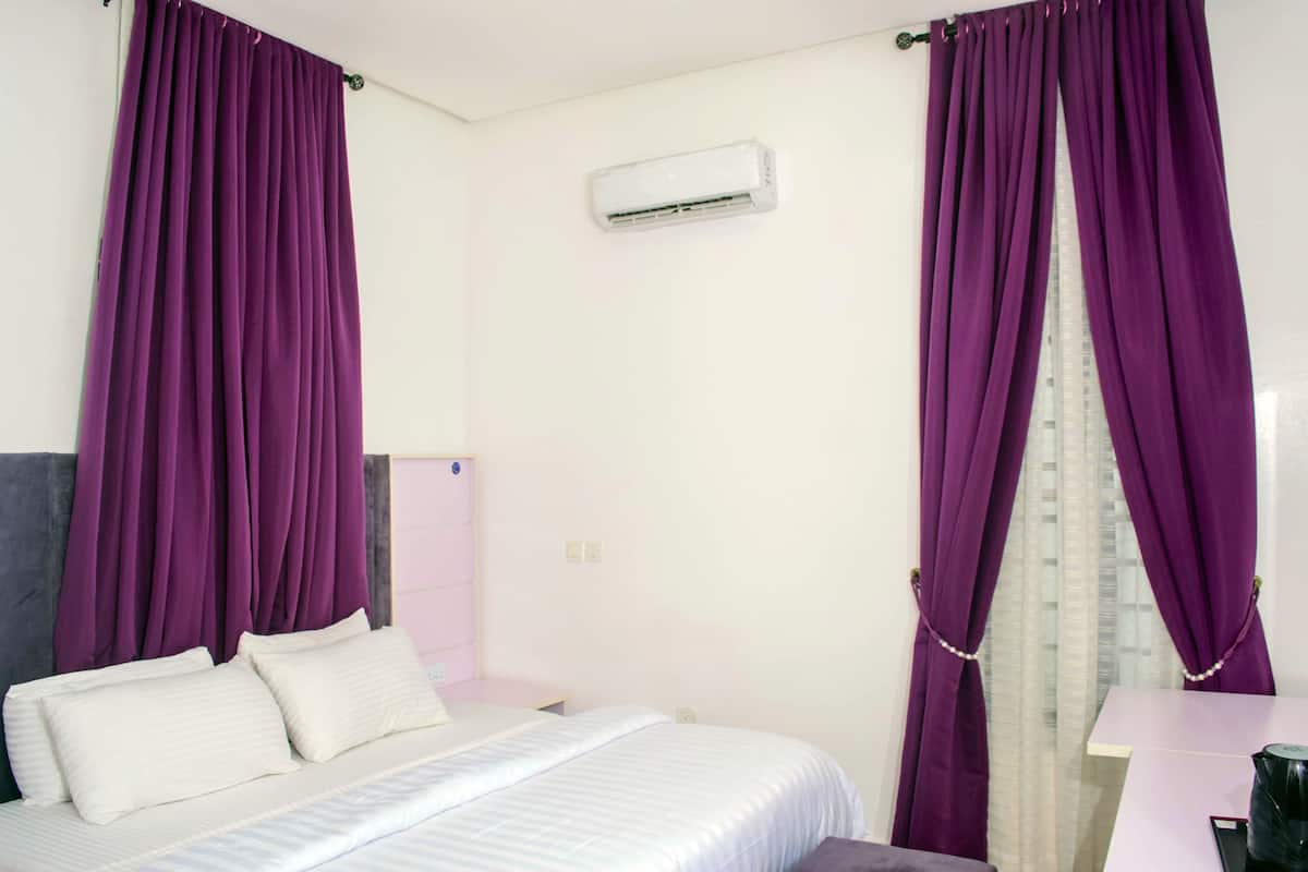 A bedroom with a bed, purple curtains, an air conditioning unit, and a window with a view of buildings.