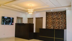 Una zona de recepción con diseño moderno, que incluye un mostrador negro, un televisor empotrado en la pared mostrando una hora, y una pared decorativa con un patrón de mosaico.