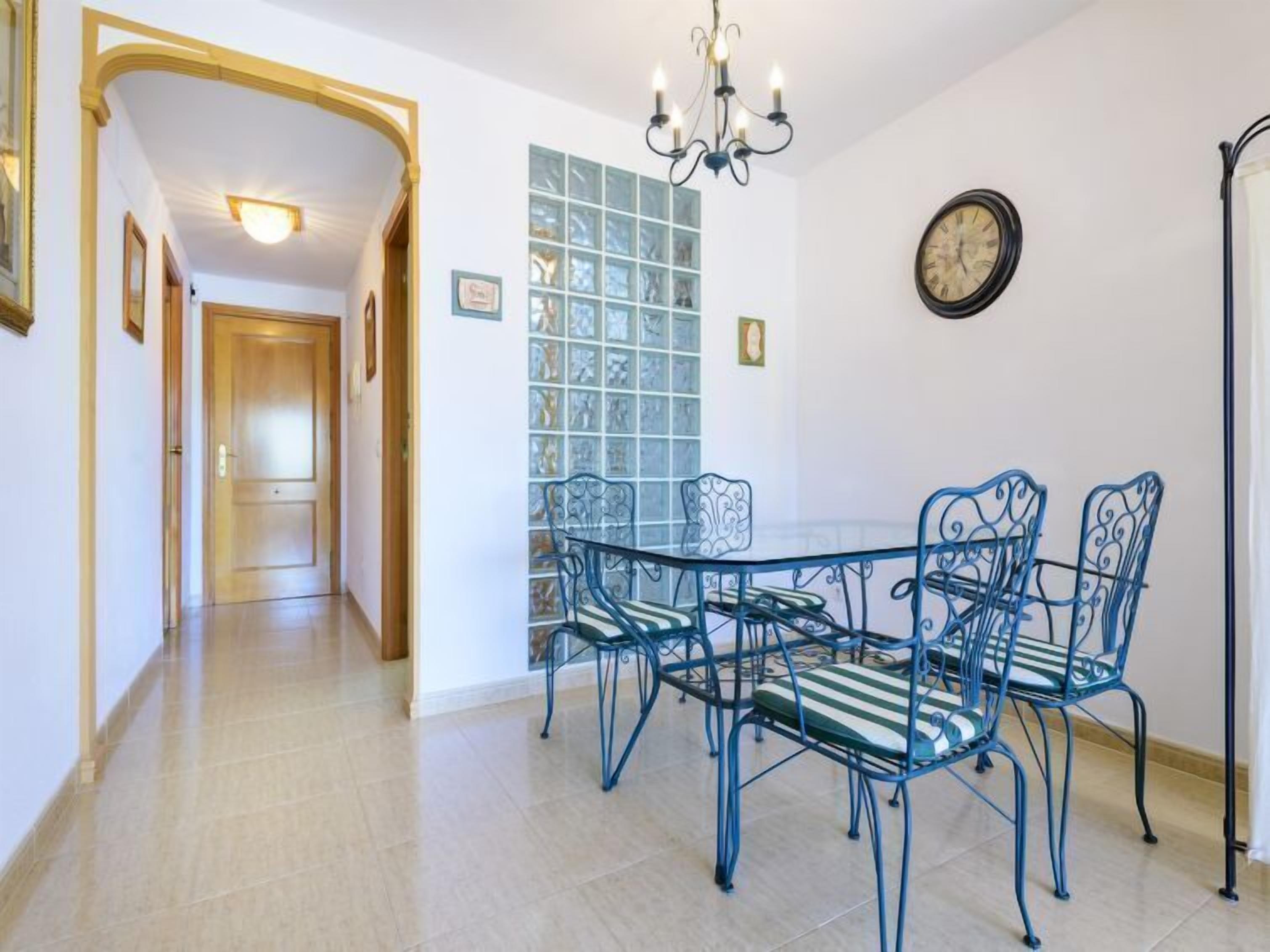 A dining area with a glass table and blue metal chairs, a chandelier, and a clock on the wall.