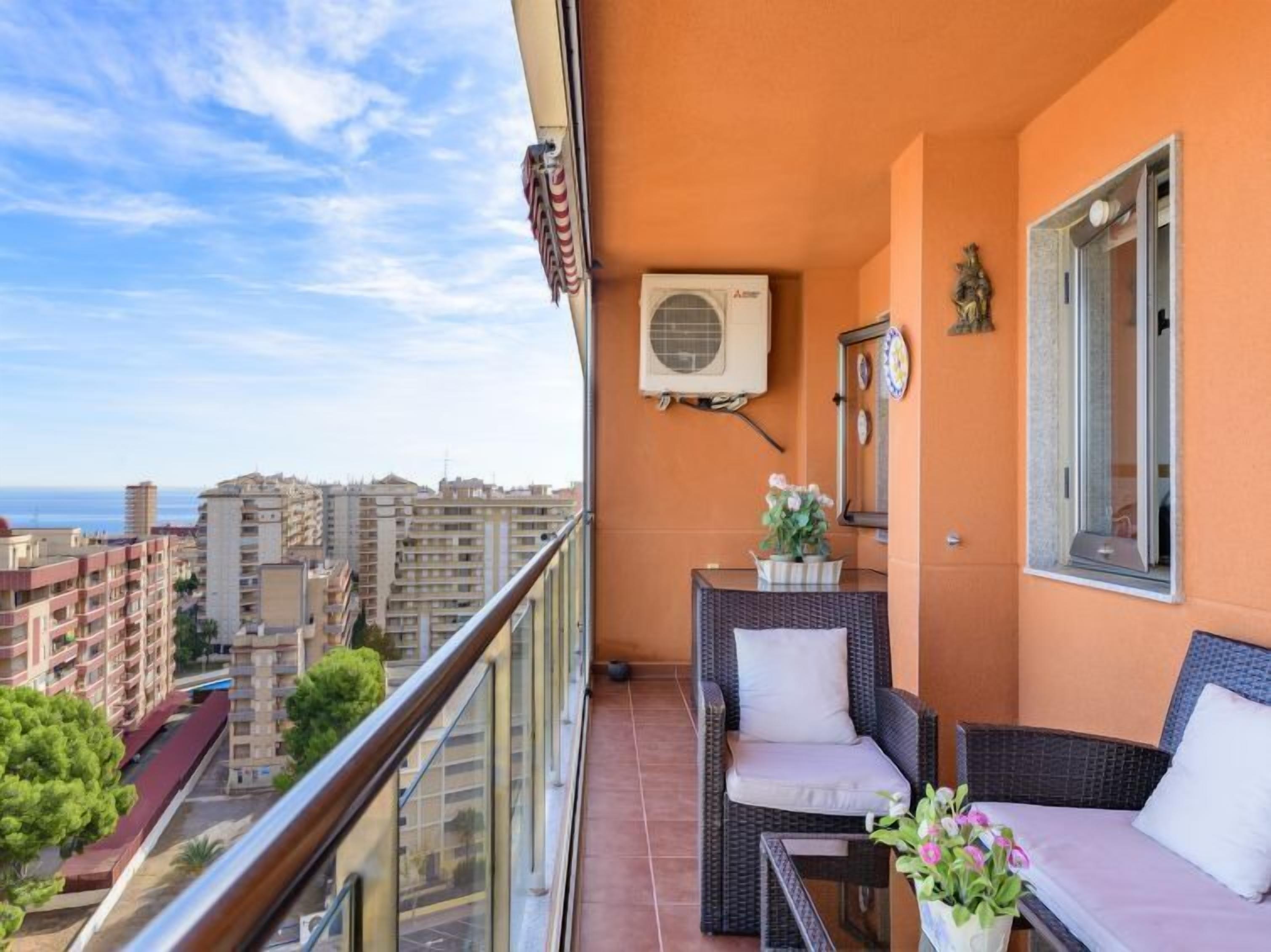 Un balcón con mobiliario de mimbre, una planta en maceta y vistas panorámicas de la ciudad.