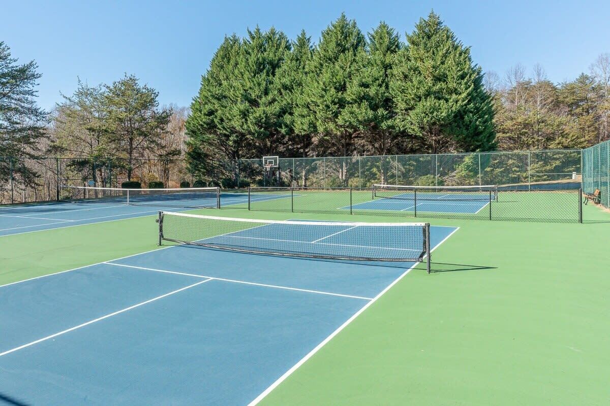 Ein Tennisplatz mit Netz in der Mitte, umgeben von einem grünen Zaun und Bäumen im Hintergrund.