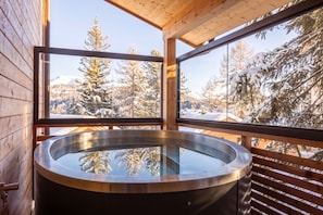 A wooden cabin with a glass-enclosed hot tub and a view of snow-covered trees and mountains.