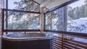 Eine Holzhütte mit großem Fenster, integriertem Whirlpool und verschneiter Aussicht.