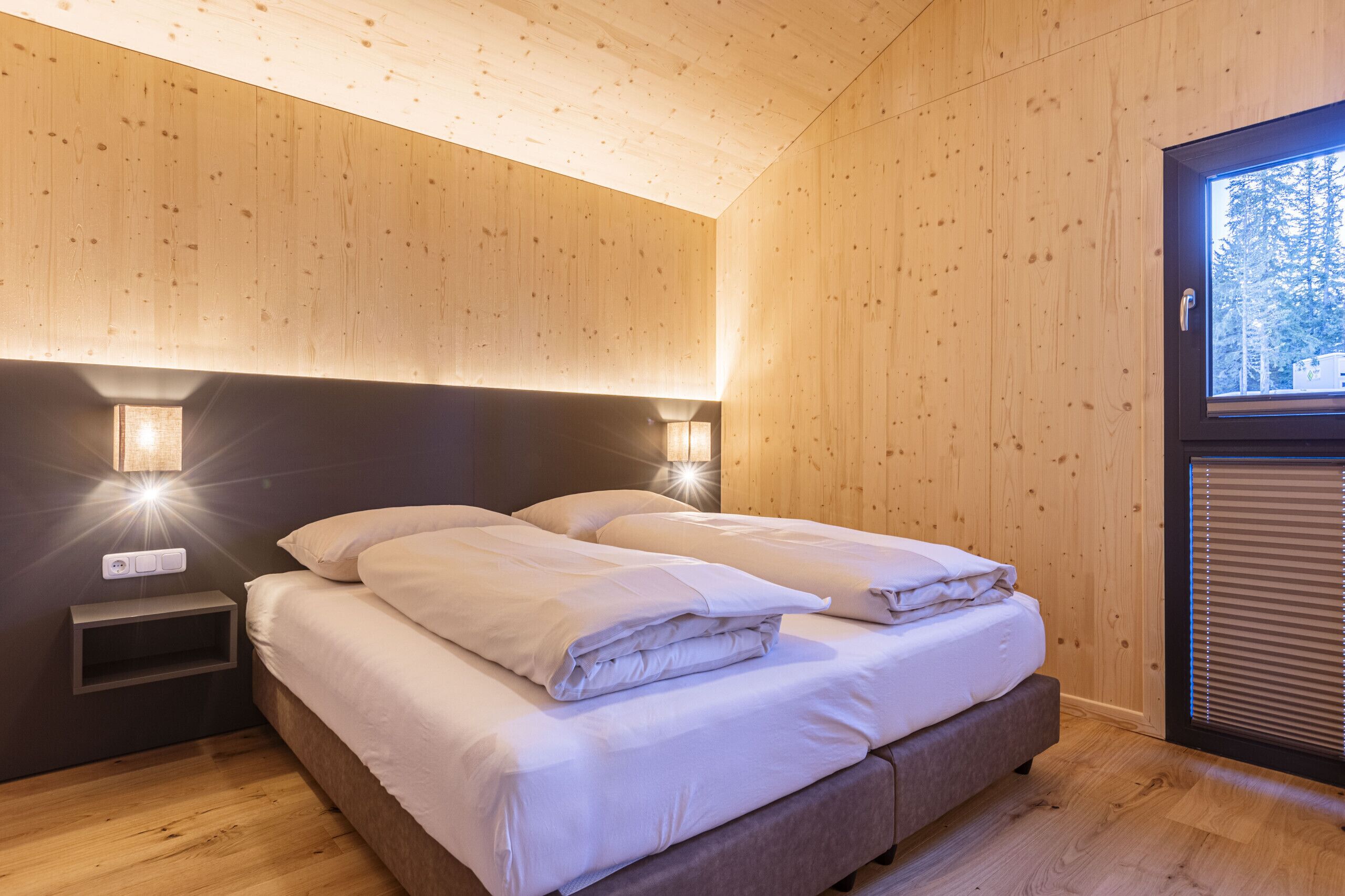 Ein modernes Schlafzimmer mit Holzboden, einem großen Bett mit weißer Bettwäsche, zwei Nachttischlampen und einem Fenster mit Jalousien.