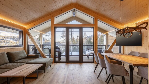 Ein geräumiger Raum mit Holzbalkendecke, einer großen Schiebeglasdoor zu einer Außenterrasse, einem Holzesstisch mit Stühlen und einem Sofa.