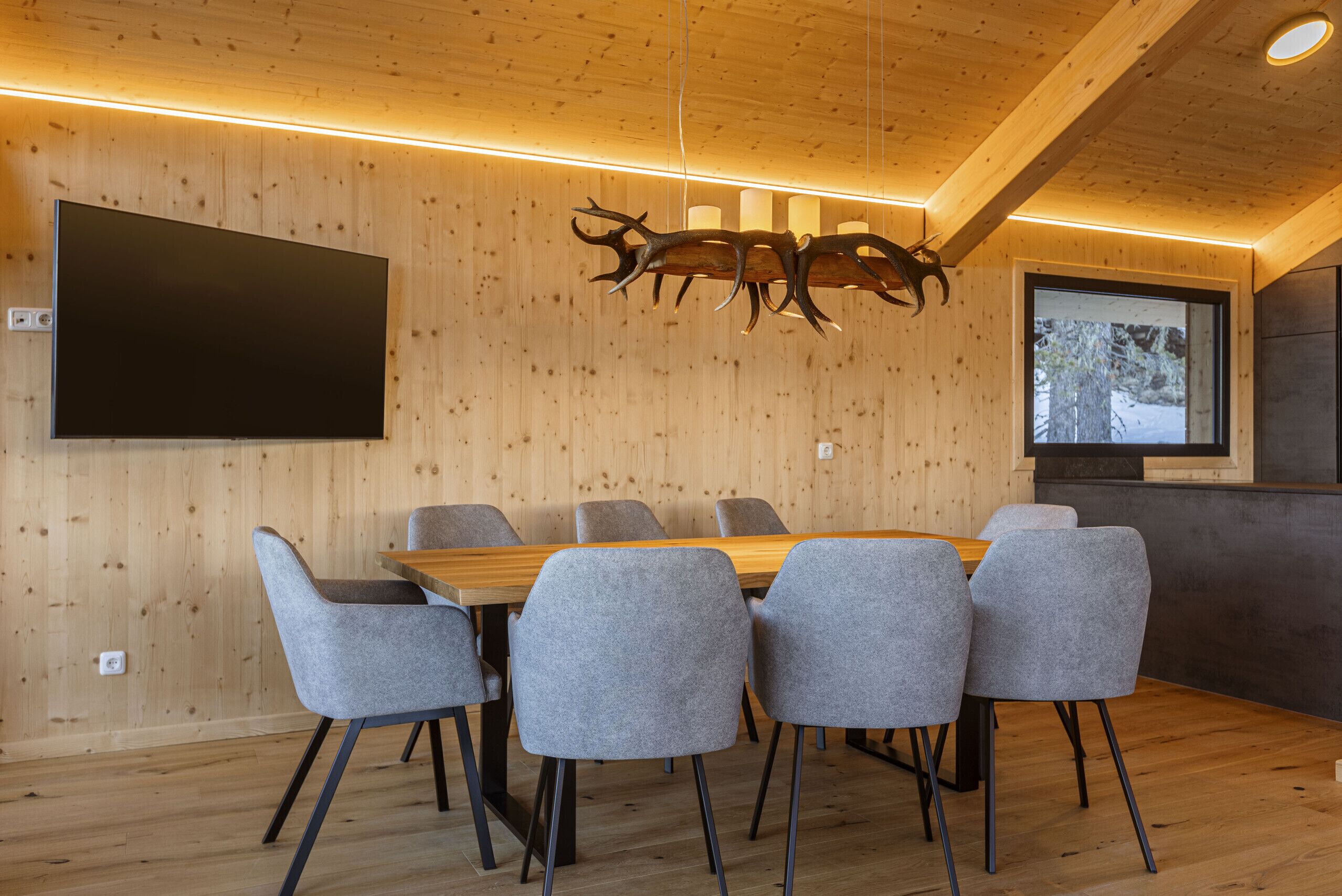 Uma sala de jantar moderna com mesa de madeira e cadeiras estofadas cinzentas, uma televisão montada e um lustre de veado original.