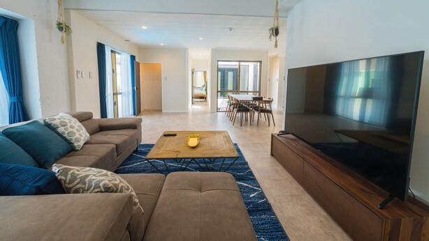 Una sala de estar moderna con sofá, mesa de centro y televisor. Se observa un área de comedor con sillas y una mesa al fondo.