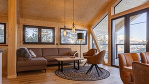 Tunnelmallinen olohuone, jossa on puinen katto, suuret ikkunat, sohva, sohvapöytä ja kaksi tuolia.