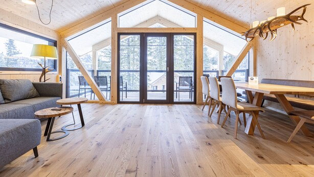 Een ruime kamer met een hoge plafond, houten balken, een grote schuifdeur naar een buitenruimte, een bank, een tafel en stoelen.