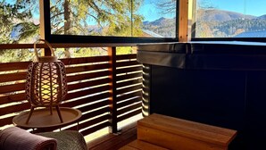 Une terrasse en bois offrant une vue sur les arbres et les montagnes, une cheminée et un coin salon avec un fauteuil en osier et des coussins.