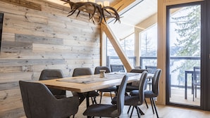 Ein moderner Essbereich mit Holztisch und -stühlen, einer Kronleuchter mit Geweihen und großen Fenstern mit Blick auf Bäume.