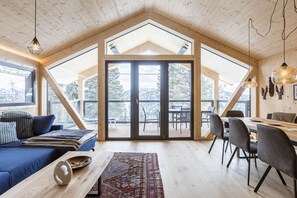 Une pièce spacieuse avec un plafond haut, des poutres en bois, une grande porte-fenêtre coulissante donnant sur une terrasse extérieure, un coin repas avec une table et des chaises, et un coin salon confortable avec un canapé et des coussins.