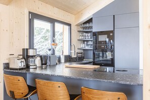 A modern kitchen with wooden cabinets, a granite countertop, and stainless steel appliances. There are two wooden chairs at the counter, and a window with a view of trees.