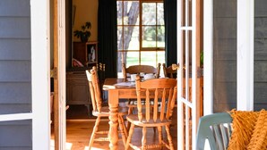 A wooden dining table with chairs, a window with curtains, and a view of greenery outside. - Ethel Cottage (Drummond)