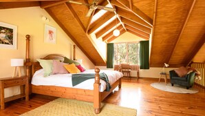 A cozy attic bedroom with a wooden ceiling, a large bed with multiple pillows, a desk, and a chair. - Ethel Cottage (Drummond)