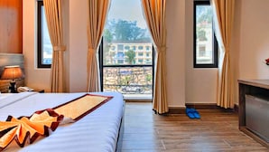 A hotel room with a bed, a nightstand, a lamp, a dresser, and a view of a building through a window. - HANZ Gia Hoang Hotel Dalat (Cam Ly - Da Lat)