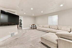 A spacious living room with a large flat-screen TV, a beige sectional sofa, and a small table with chairs. - New 4BR Nephi, UT Home - 12 Beds, Game Garage, RV Pad, Near Mt. Nebo & I-15 (Nephi)
