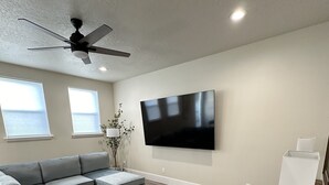 Una sala moderna con un sofá seccional, un televisor de pantalla plana, un ventilador de techo y una mesa de centro de madera.