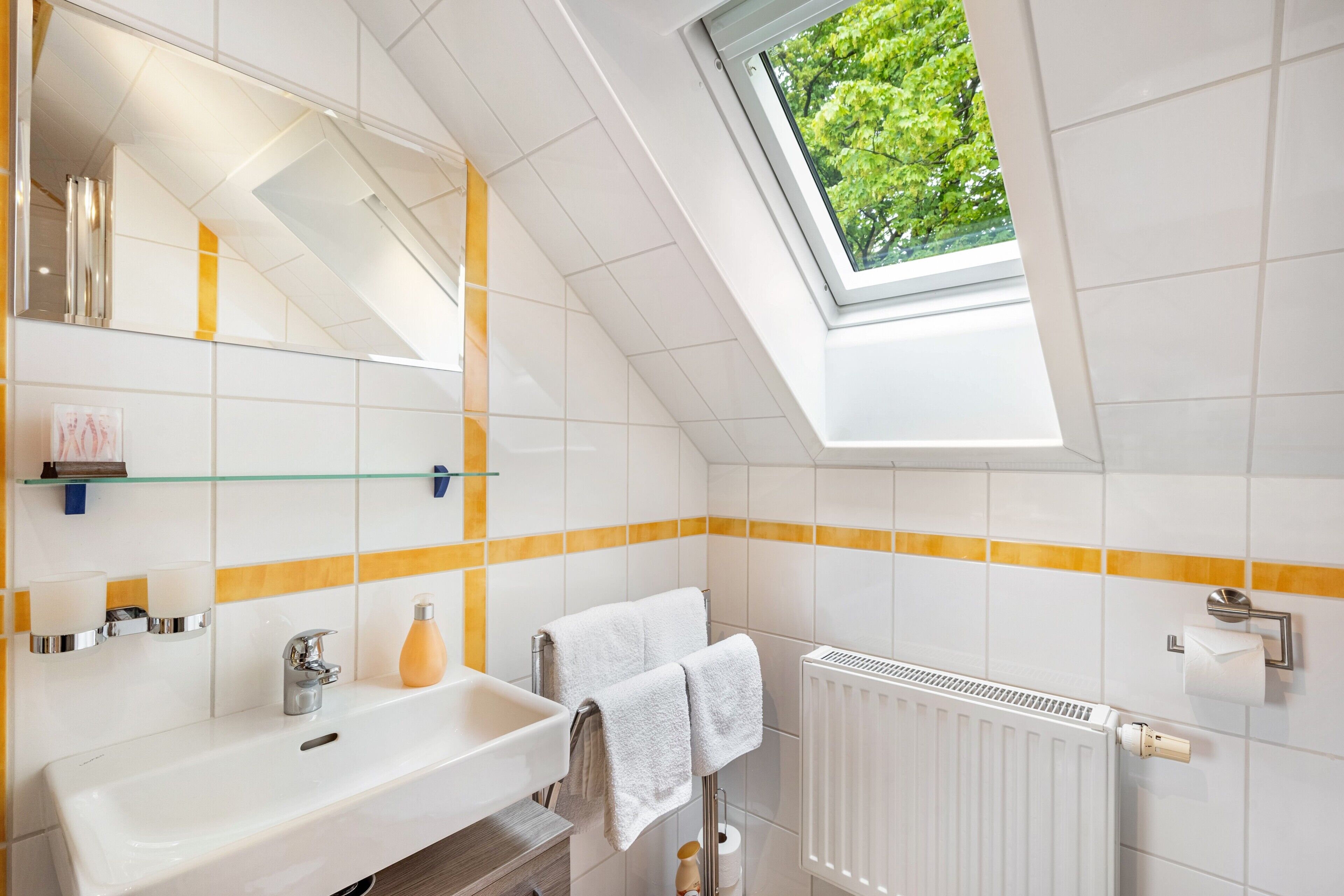 Een moderne badkamer met dakraam, witte tegels, een glazen plank, een wastafel, een handdoekhouder en een toiletrolhouder.