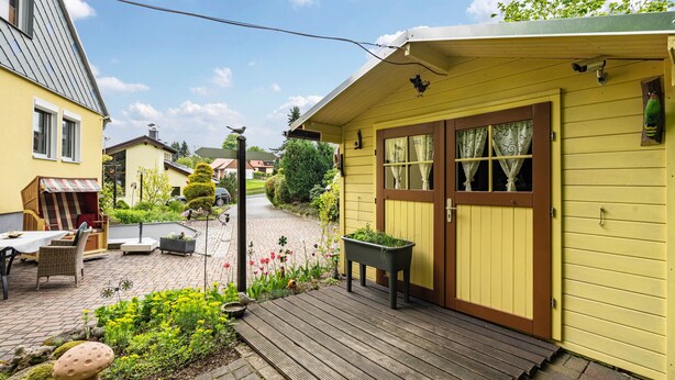 A yellow house with a wooden deck, a garden with flowers and a mushroom, and a neighboring building. - Apartment 'Ferienwohnung Lucke' with Mountain View, Private Terrace and Wi-Fi (Oybin-Luftkurort Lückendorf)
