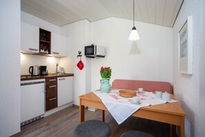 A compact kitchen with a microwave, kettle, and coffee maker. A dining table with a red and white checkered tablecloth and a vase of red flowers is set for two.