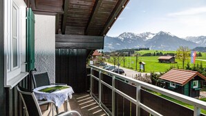 Un balcon avec une table et des chaises, une vue sur une chaîne de montagnes et un petit bâtiment au loin.
