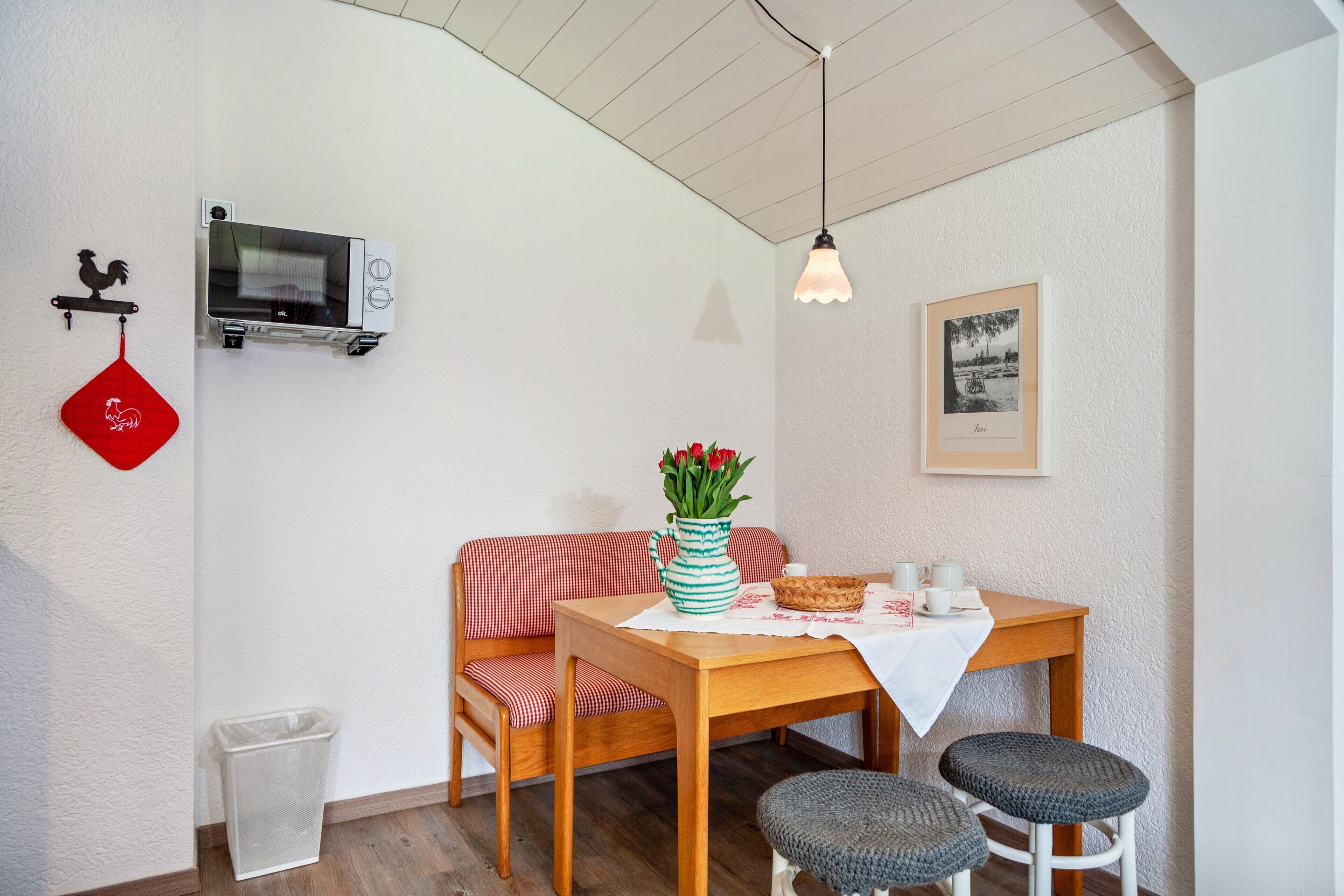 A dining area with a wooden table, two chairs, a red and white checkered sofa, a vase with flowers, a microwave, a trash bin, and a framed picture on the wall.