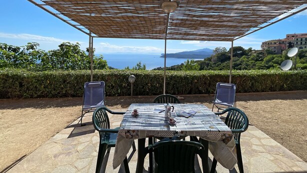 An outdoor dining area with a table and chairs, a pergola overhead, and a view of the sea and hills.
