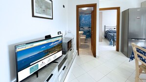 A modern kitchen with a refrigerator, a dining table, and a TV displaying a travel website. A bathroom with a shower and a bed is visible through an open door.