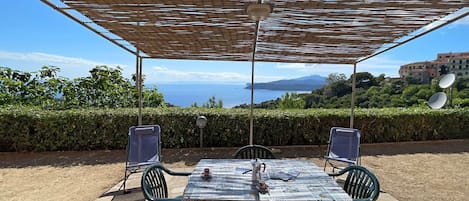 An outdoor dining area with a table and chairs, a pergola overhead, and a view of the sea and hills.