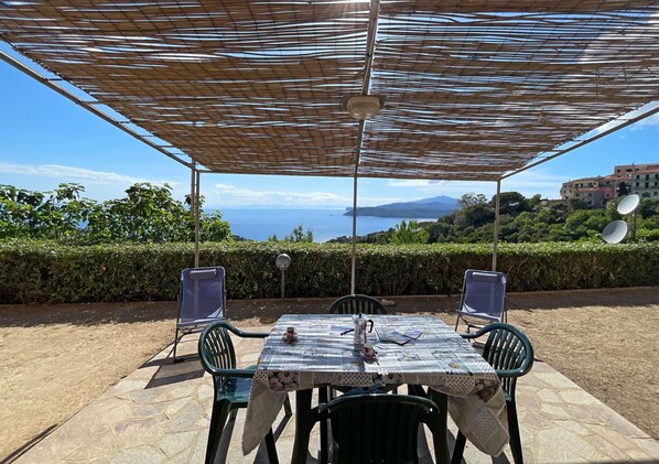 Un espacio para comer al aire libre con mesa y sillas, una pérgola arriba y vistas al mar y las colinas.